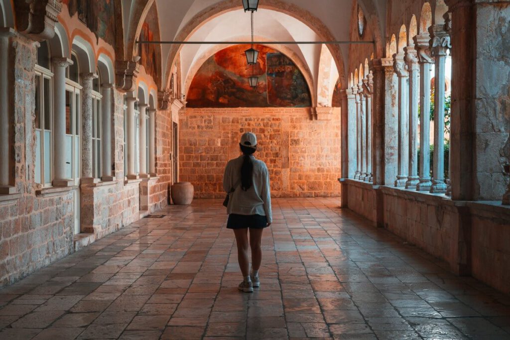 Frau läuft durch Gänge des Franziskaner Klosters und bestaunt die bogenförmigen Wandmalereien mit Szenen aus dem Leben des heiligen Franziskus von Assisi